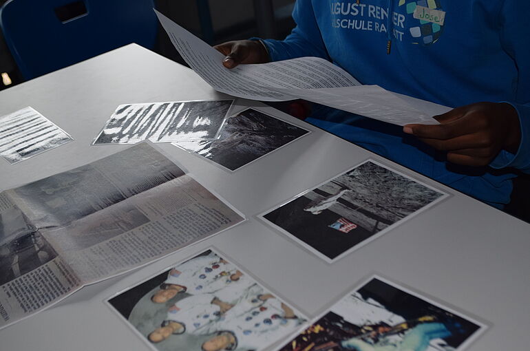 Das Bild ist auf den Tisch ausgerichtet und man sieht verschiedene Bilder und Artikel und ein Junge (abgeschnitten) der ein Blatt in der Hand hält