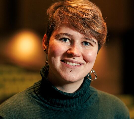 Junge Frau mit kurzen braunen Haaren und einem grünen Rollkragenpulli lächelt in die Kamera.