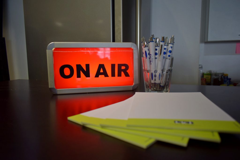 eine rotblinkende Onair Lampe, die sagt hier wird Radio gemacht. Daneben viele Stifte und Blöcke mit LKJ Logo, die bereit stehen, um damit zu arbeiten.