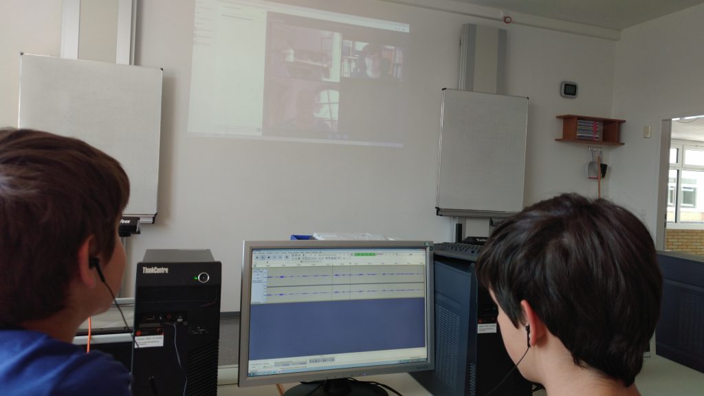 Bild: Zwi Teilnehmer*innen sitzen vor ihrem Schnittprogramm am Computer auf der Leinwand davor sieht man die beiden referenten in der Videokonferenz