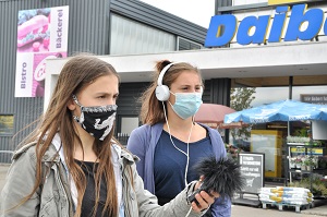 Zwei Teilnehmerin stehen mit Aufnahmegerät und Kopfhörern vor einem Einkaufszentrum bereit, um Stimmen für ihre Umfrage einzusammeln