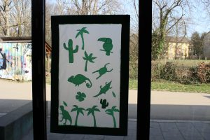 An einem Fenster wurde papier aufgehängt auf welchem grüne Pflanzen und Tiere zusehen ist.
