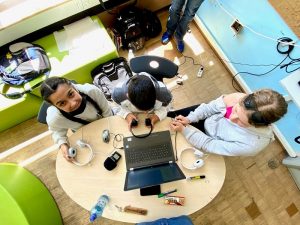 drei Jugendliche sitzen an einem Tisch voller Technik. Ein Laptop, viele Kopfhörer, ein Aufnahmegerät und um sie herum ganz viele Kabel. 