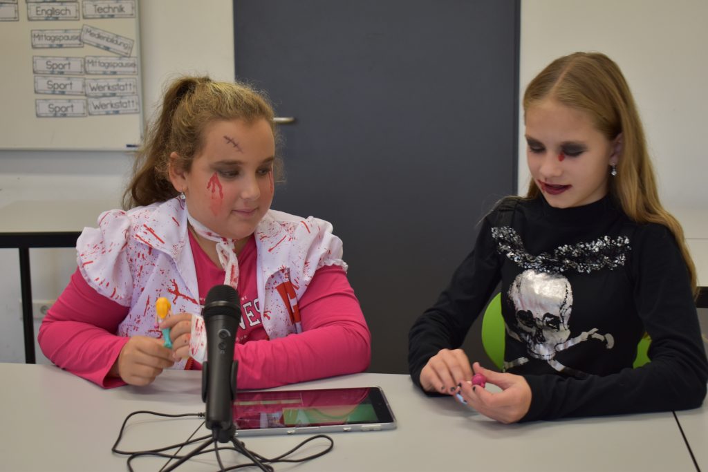 Zwei gruselig verkleidete Kinder sitzen am Tisch und machen Tonaufnahmen mit einem Mikrofin vor ihnen.