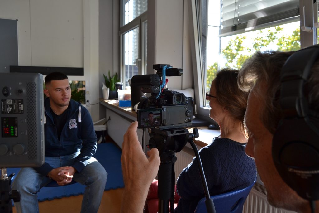 Auf dem Foto sitzt ein Jugendlicher vor einer Kamera, welche von einem Mann bedient wird. Zusätzlich ist noch eine Frau im Bild zu sehen, die den Jugendlichen interviewt.
