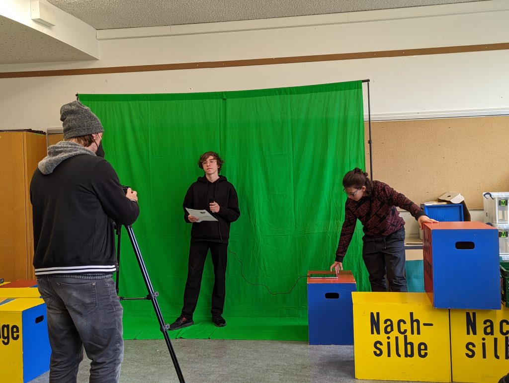 Auf dem Foto sind zwei Schüler vor einem großen grünen Stoffvorhang zu sehen. Ein Medienreferent filmt die Szene.