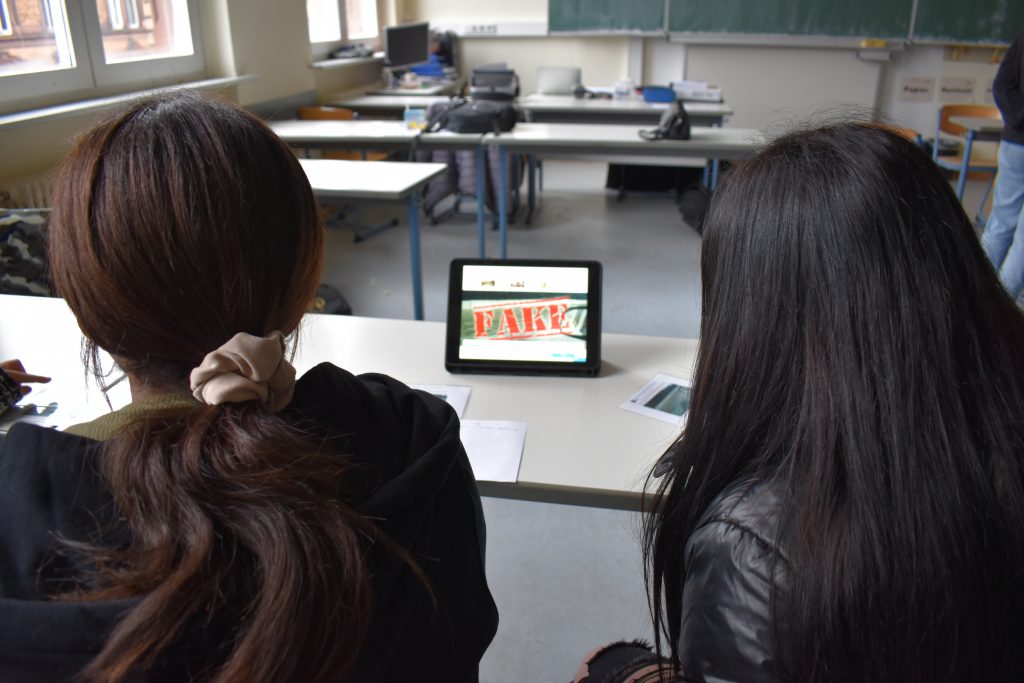 Auf dem Foto sind zwei Schülerinnen von hinten zu sehen, die auf ein Tablet schauen. Auf dem Tablet steht "Fake".