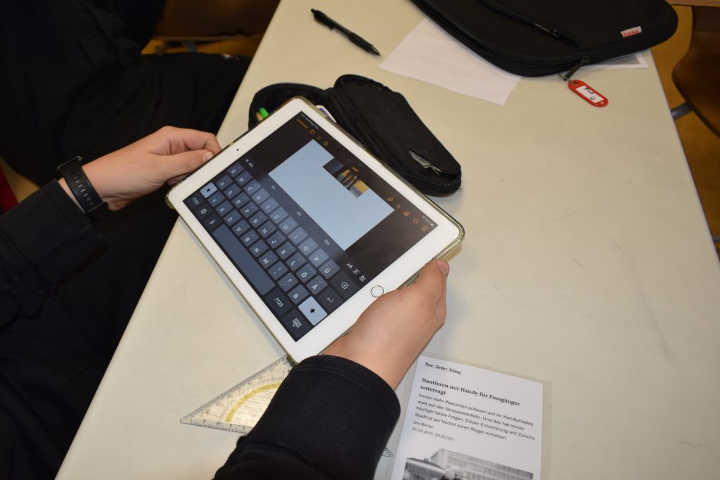 Auf dem Foto ist ein iPad zu sehen und zwei Hände, die dieses bedienen. Außerdem befindet sich ein ein Mäppchen und ein Zeitungsausschnitt mit im Bild.