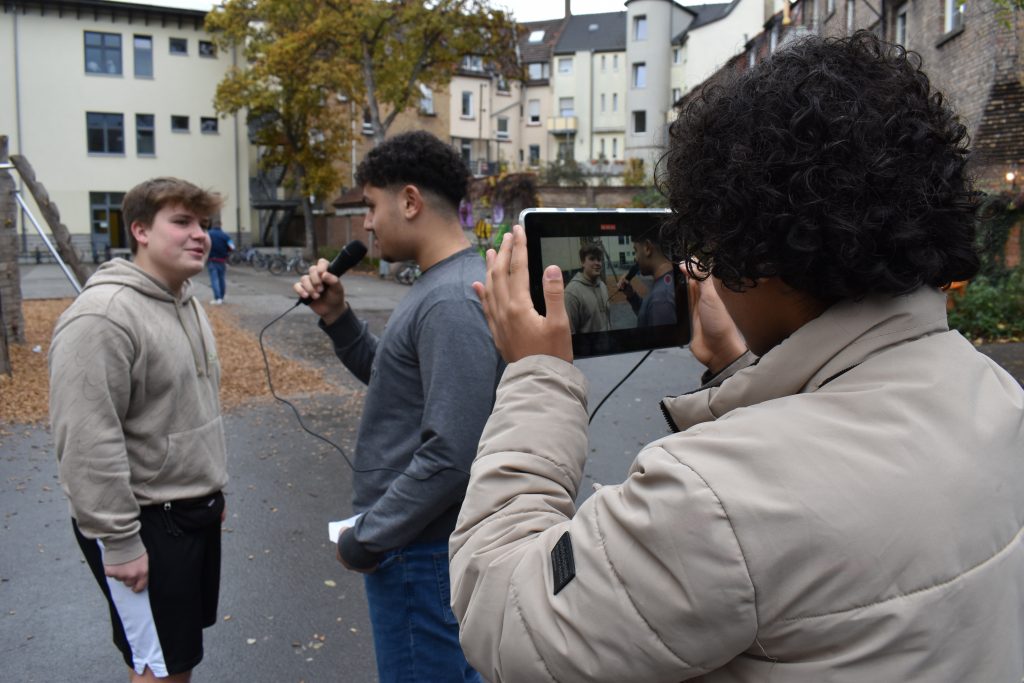 Drei Jungs sind auf dem Pausenhof einer Schule. Ein Junge filmt, während die anderen beiden ein Gespräch mit Mikrofon führen.