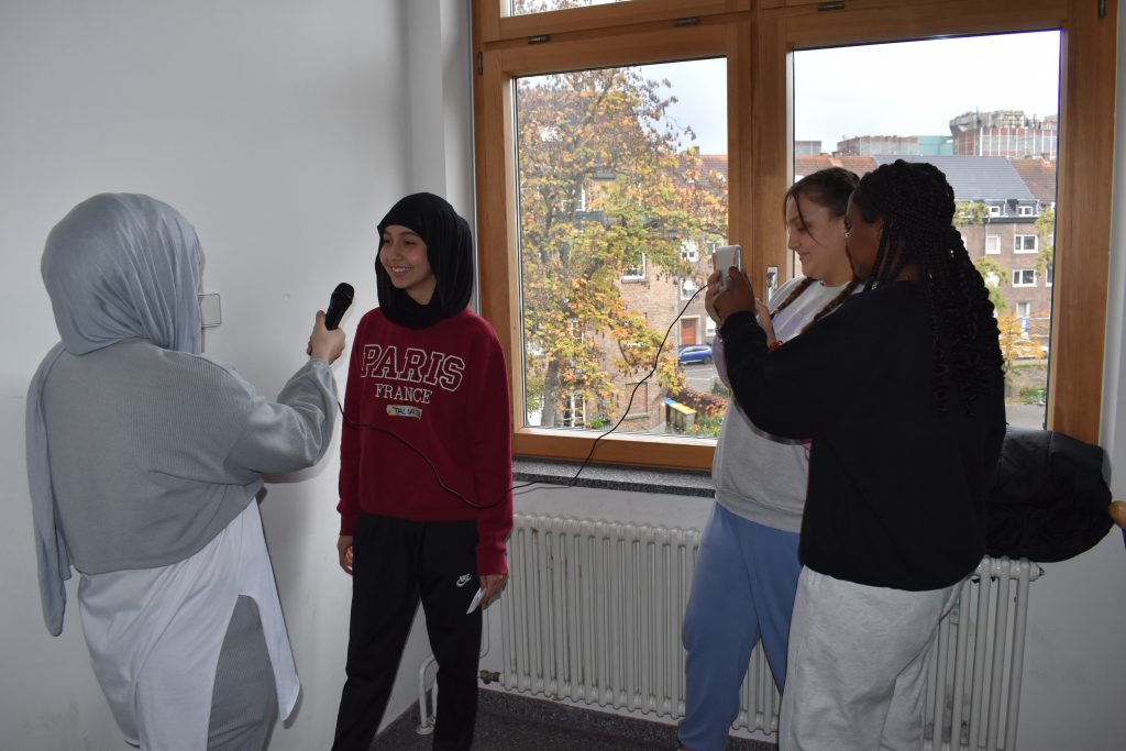 Vier Mädchen (zwei davon mit Kopftuch) stehen in einem Raum vor einem fenster. Zwei Mädchen Filmen mit einem Tablet, die anderen zwei Mädchen befinden sich in einer Interviewsituation.