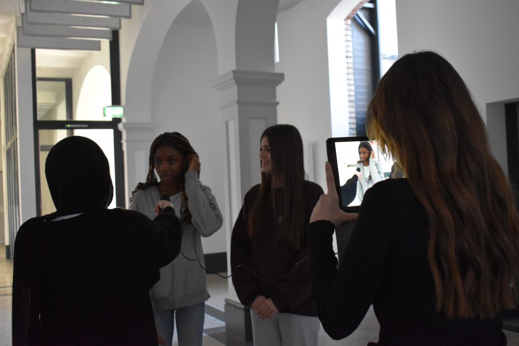 Vier Schülerinnen stehen im Flur einer Schule und filmen sich während eines Interviews mit dem Tablet.