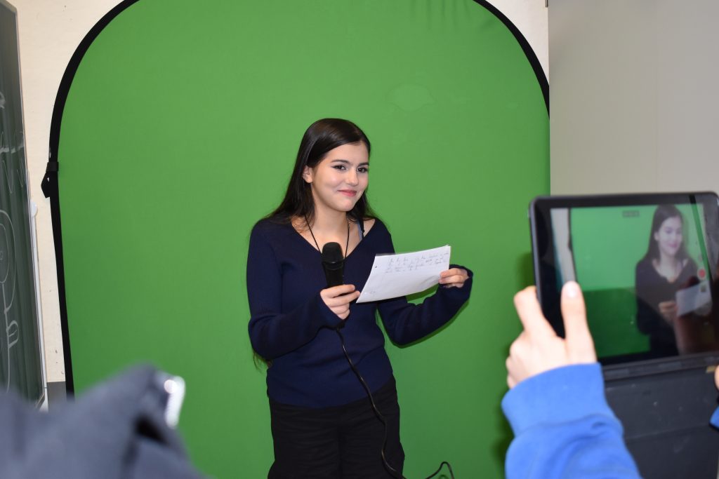 Auf dem Foto ist eine Schülerin vor einem Greenscreen zu sehen, wie sie ein Mikrofon in der Hand hält und gefilmt wird.