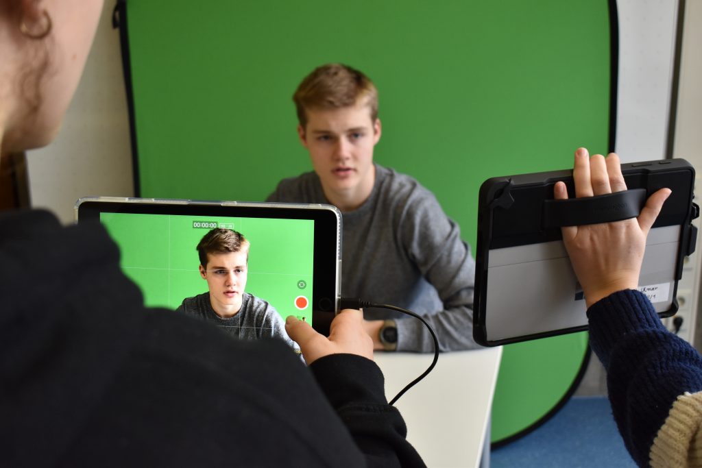 Ein Schüler sitzt vor einem Greenscreen und wird mit einem Tablet gefilmt.