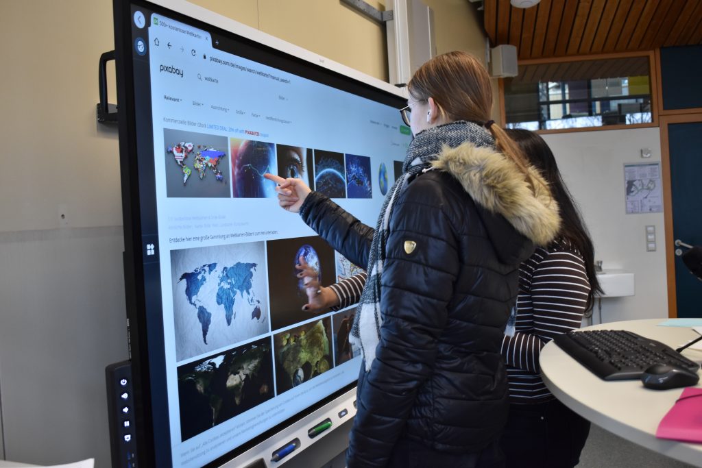 Zwei Schülerinnen stehen vor einem großen Smartboard im Klassenzimmer und suchen im Internet Bilder.