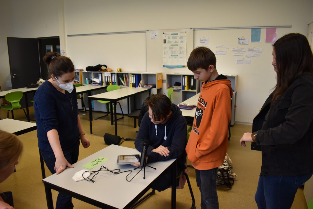 Zwei Schüler und eine Schülerin stehen um einen Tisch im Klassenzimmer. Vor ihnen steht ein iPad und ein Mikrofon. Eine Medienreferentin erklärt die Technik.