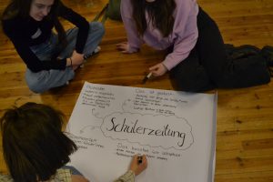 Drei Mädchen sitzen auf dem Bofen und schreiben auf ein Plakat. Überschrift auf dem Plakt ist Schülerzeitung