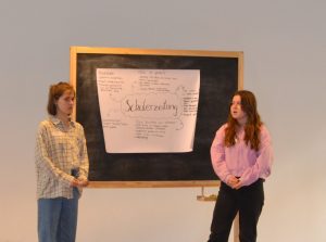 Zwi Mädchen stehen vor einer kleinen Tafel auf der ein Plakt ist