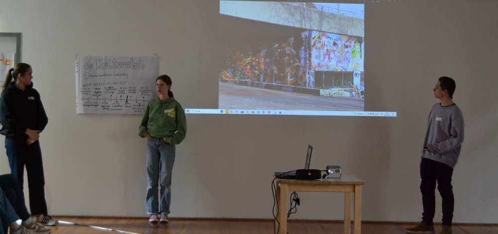 3 Jugendliche präentieren ihr Kulturprojekt mit einem Plakat und einer Powerpointpräsentation
