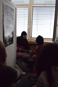 Blick in einen kleinen Raum, in dem ein Mädchen an der Gitarre sitzt und wohl gerade ihren eigenen Song aufnimmt. Publikum hat sie auch um sich herum.