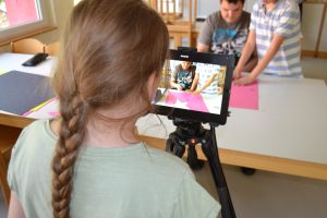Eine Schülerin bedient ein Tablet und filmt zwei weitere Schüler beim Basteln.
