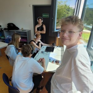 Schüler*innen sitzen um einen Laptop, mit dem ihre Kopfhörer Verbunden sind. Manche hören sichtlich konzentriert zu andere posen sympatisch für die Kamera.
