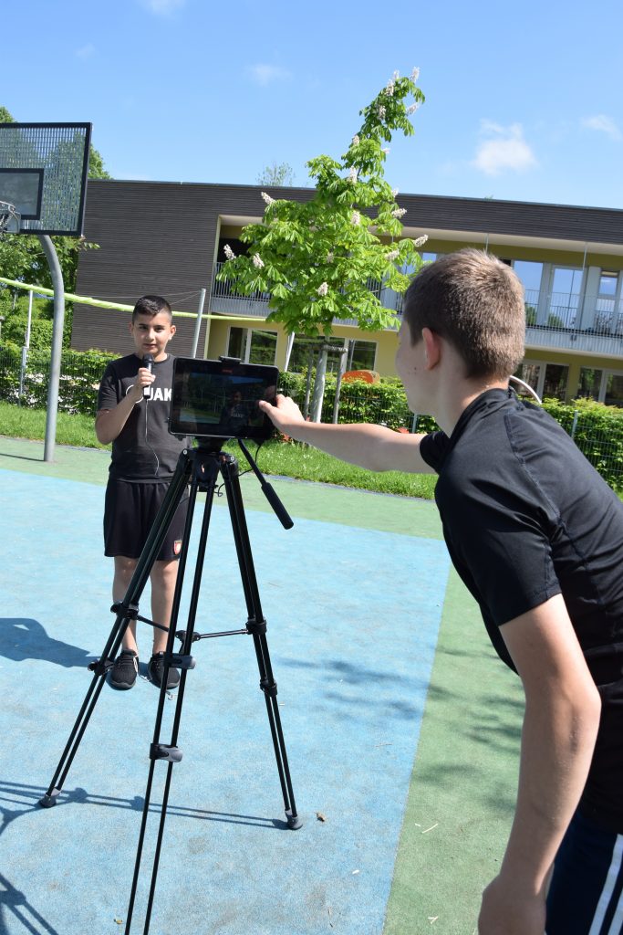 Zwei Schüler befinden sich auf einem Sportplatz. Ein Schüler moderiert mit Mikrofon, ein anderer filmt mit Stativ und Tablet.