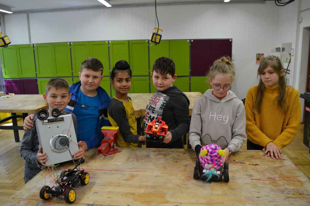 Sechs Kinder präsentieren vier fertige Kunstroboter um einen Tisch stehend.