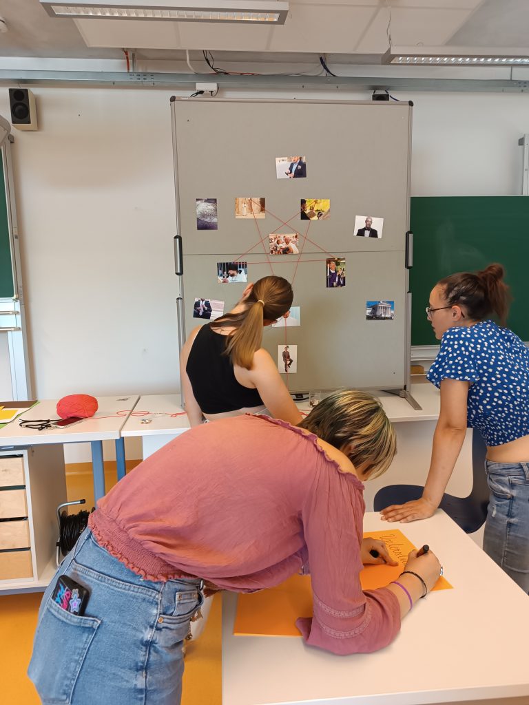 Drei Schülerinnen stehen vor einer Pinnwand, welche mit Fäden und Fotos bestückt ist.