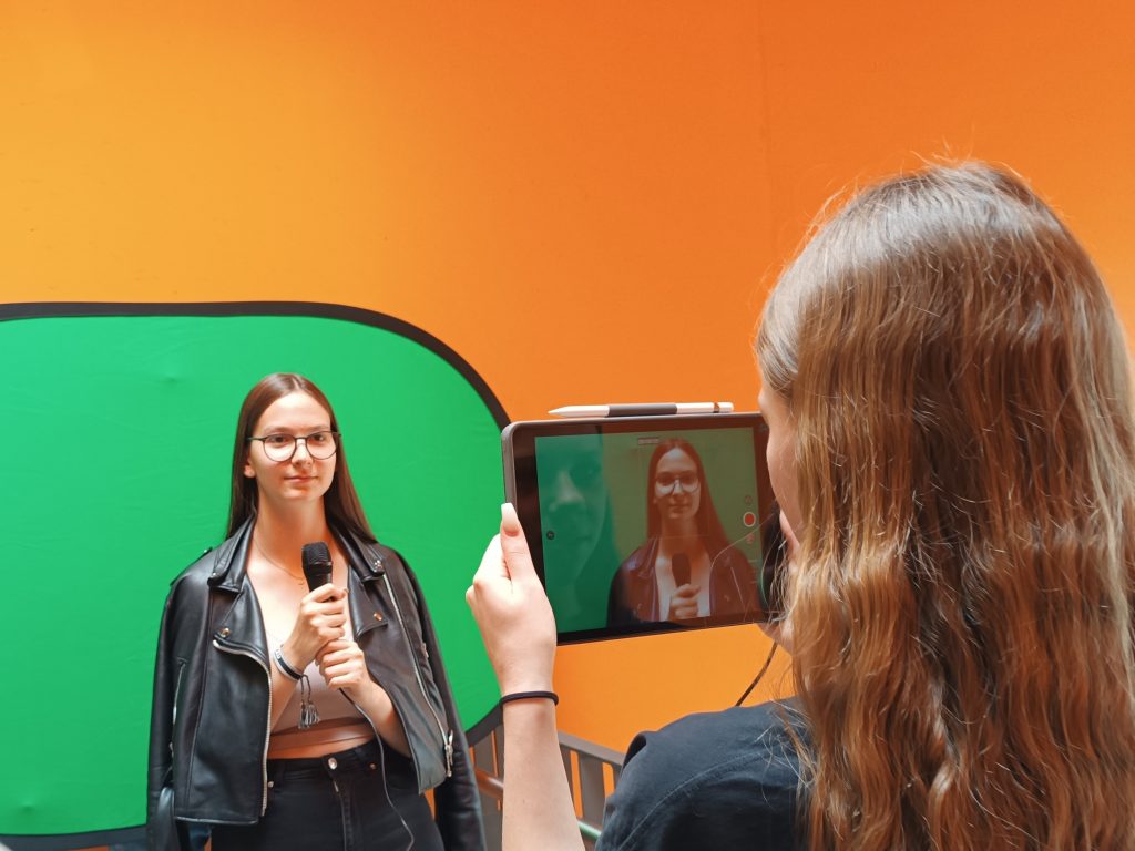 Eine Schülerin steht vor einem Greenscreen und wird mit einem Mikrofon in der Hand von einer Mitschülerin gefilmt.