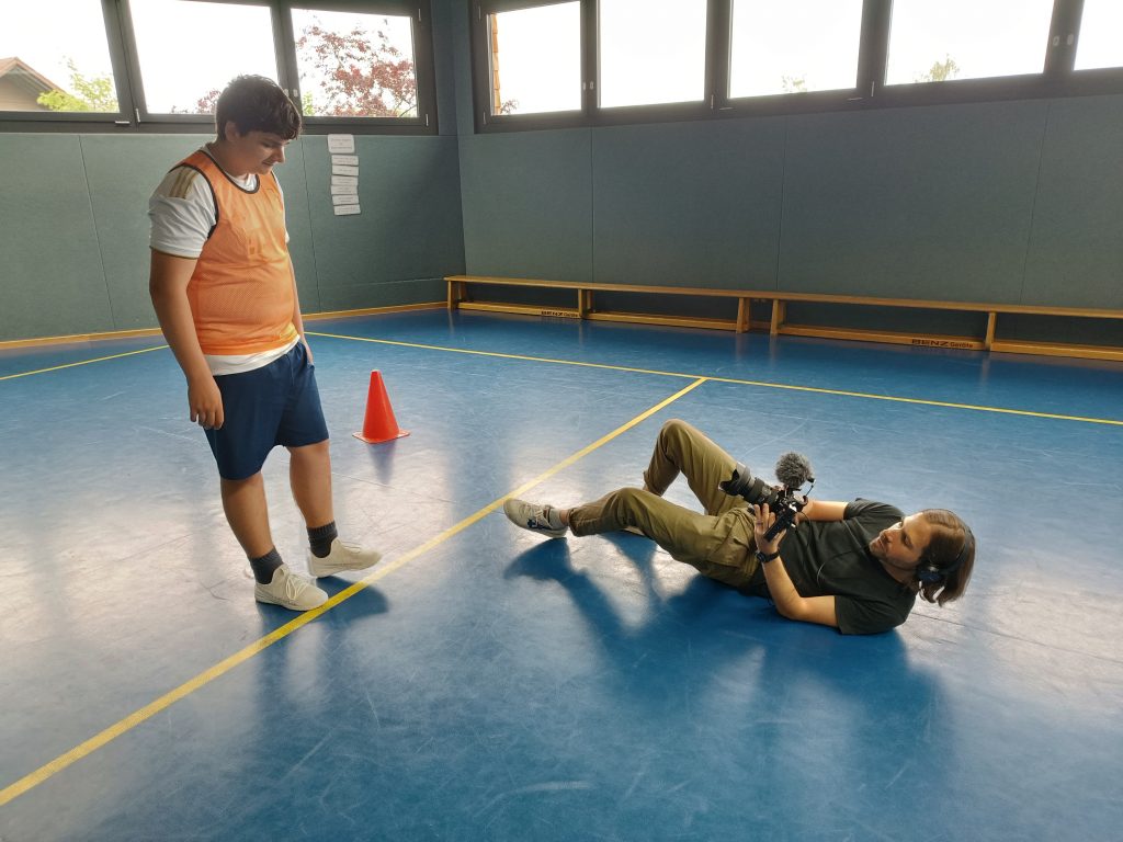 Ein Schüler befindet sich in einer Sporthalle und wird von einem Mann, der auf dem Boden liegt gefilmt.