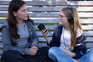 ein Mädchen mit Aufnahmegerät und Kopfhörern hört aufmerksam der Antwort Ihrer Interviewpartnerin zu