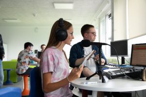 Eine Schülerin vor dem Mikrofon der Live Sendung. Im Hintergrund applaudiert der Workshopleiter sichtlich stolz