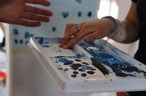 Ein Finger taucht in blaue Farbe, im Hintergrund sieht man die Wand, die bereits einige Fingerabdrücke bekommen hat.