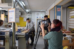 junger Mann arbeitet mit Holz, trägt Gehörschutz. Dabei wird er von einer Frau mit einem Tablet gefilmt.