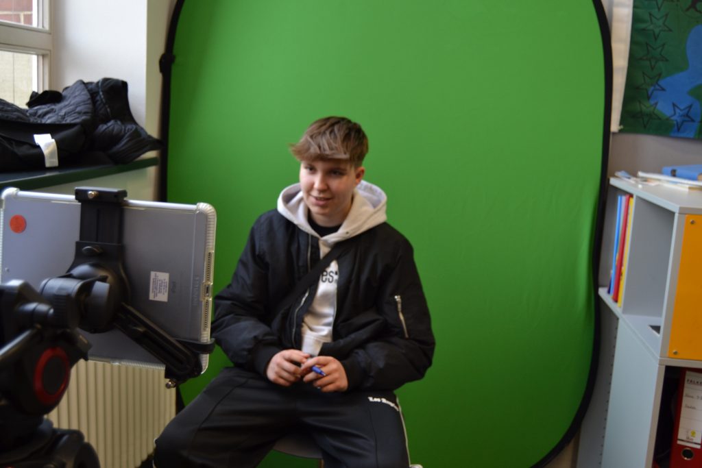 Jugendlicher sitzt auf einem Stuhl vor Greenscreen und schaut in eine Kamera. 