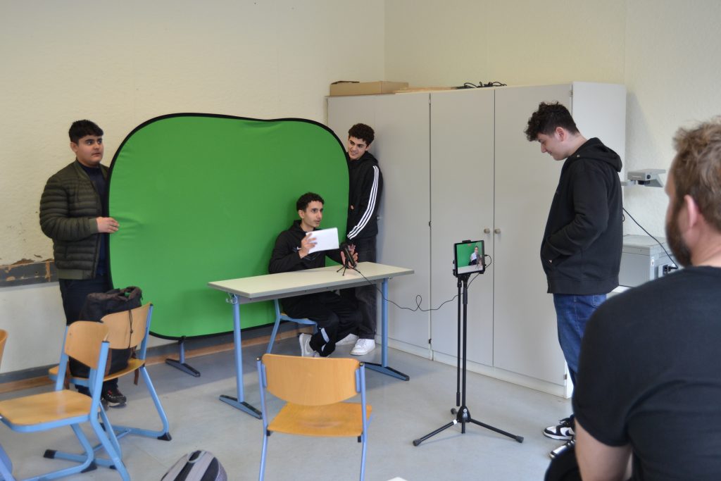 Jugendlicher sitzt an einem Tisch mit einem Mikrofon vor sich. Hinter ihm halten zwei Jugendliche einen Greenscreen, er wird mit einem Tablet gefilmt.