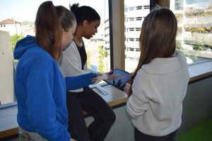 Drei Jugendliche stehen/sitzen vor einer Fensterfront und schauen auf ein Tablet, zeigen mit den Fingern darauf.