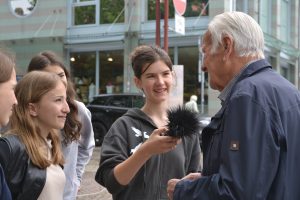 Vier Schülerinnen stehen mit einem Mann mit weißem Haar auf der Straße gegenüber und schauen ihn freundlich an. Eine von ihnen Hält dem Mann ein Aufnahmegerät entgegen in das er zu spricht.