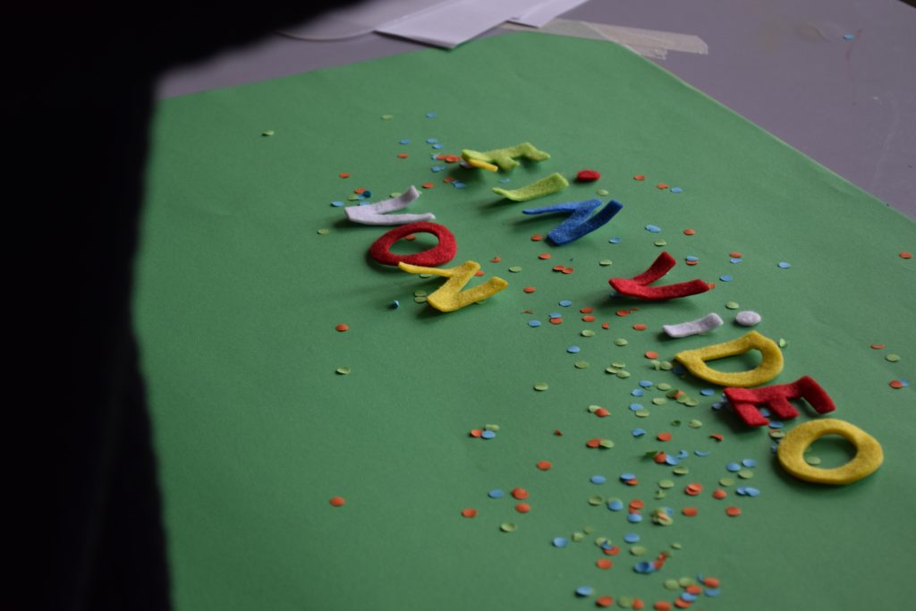 Auf grünem Untergrund liegen Filz-Buchstaben. "Ein Video von" ist zu lesen. Drumherum liegt Konfetti.