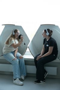 zwei Mädchen mit Kopfhörern sitzen sich gegenüber. Eine der beiden spricht in ein Aufnehmegerät