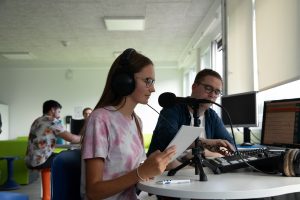 Eine junge Teilnehmende spricht in ein Mikrofon und liest von einem Blatt ab. Neben ihr dreht ein Workshopleiter an verschiedenen Reglern.