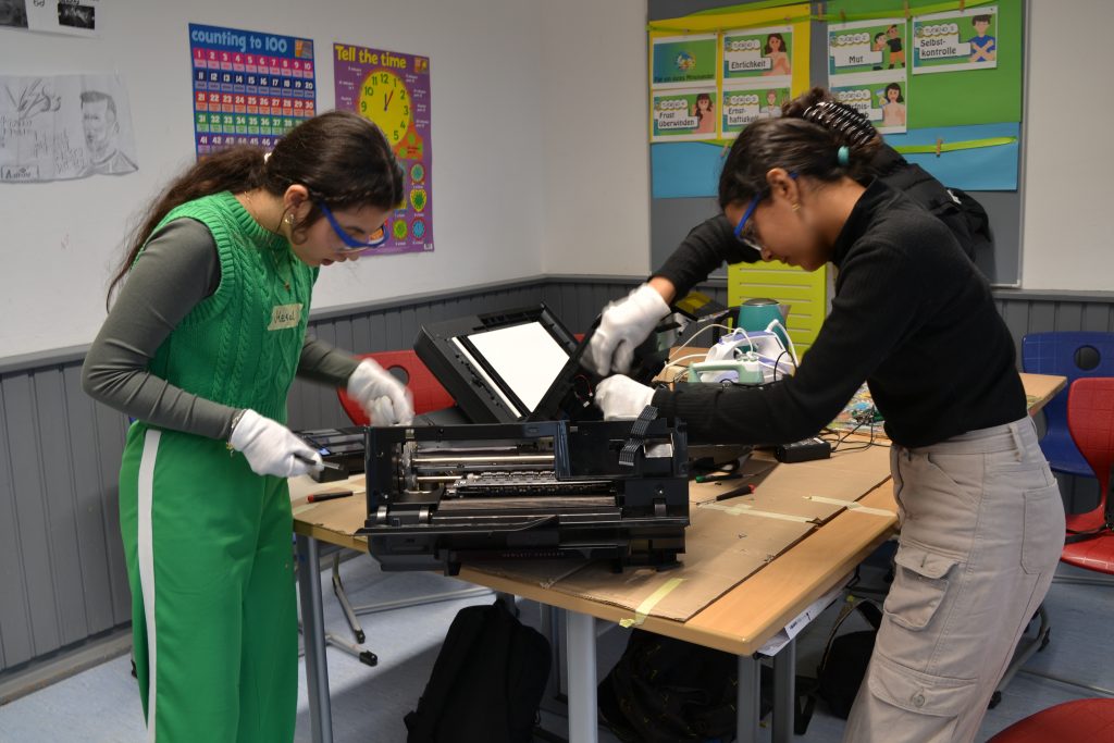 zwei Jugendliche stehen mit Schutzbrillen und Handschuhen über einem auseinandergebauten Gerät und schrauben daran.