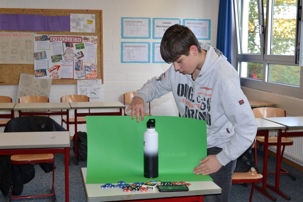 Jugendlicher hat auf einem Tisch eine Trinkflasche, Casino-Chips, ein kartenspiel und ein Handy verteilt und hält ein grünes Papier hinter die Flasche.