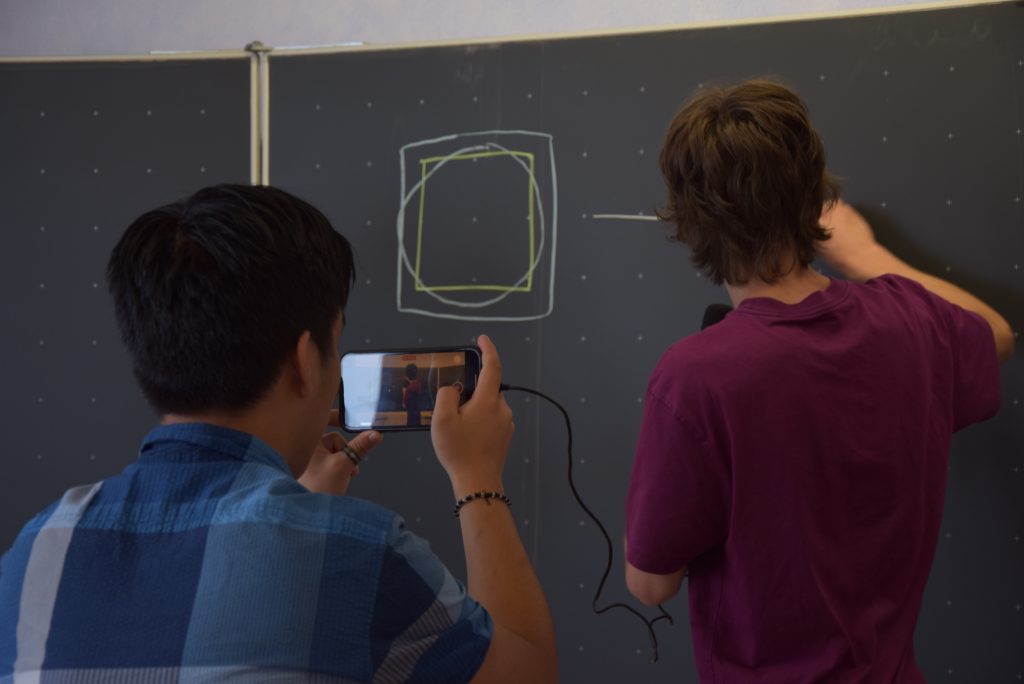 Schüler malt eine Grafik an die Tafel, ein anderer filmt ihn dabei.