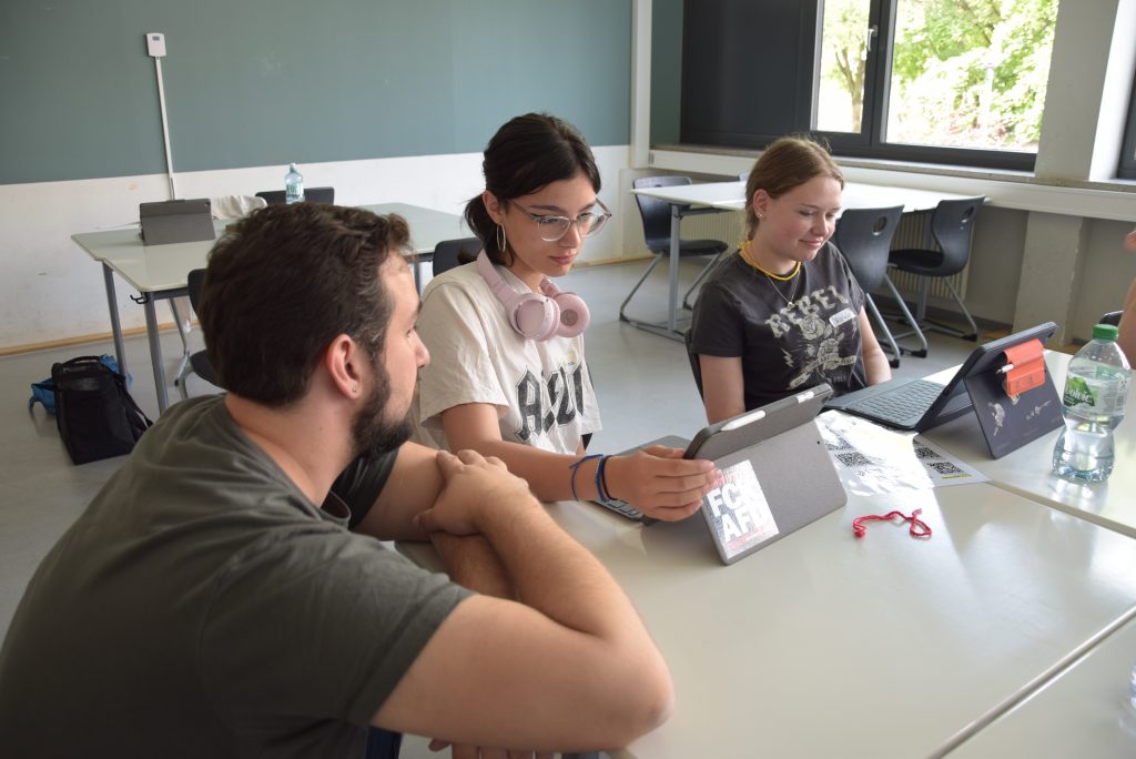 zwei Jugendliche sitzen an Schultischen nebeneinander, Tablets vor Ihnen. Ein Referent kniet daneben und lehnt sich auf den Tisch.