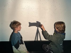 Zwei jungen stehen sich gegenüber zwischen ihnen der Schatten einer Kamera und der rechte jungen hebt die hand sodass es aussieht, er würde den anderen damit fotografieren.