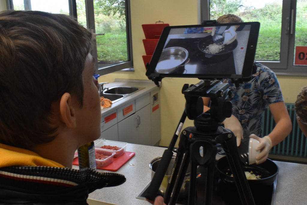 Ein Jugendlicher steht vor einem Tablet, das auf einem Stativ steht. Auf dem Display sieht man Aufnahmen, die einen anderen Jugendlichen beim Kochen zeigen.
