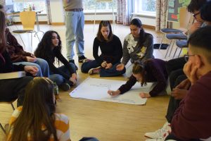 eine Gruppe sitzt zur Ideenfindung um ein Plakat auf dem eine Schülerin Ideen aufschreibt