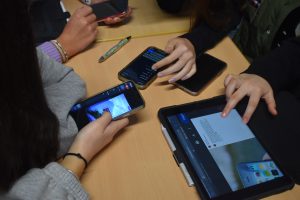 auf einem Tisch liegen drei Handys und ein Tablet. Man sieht die Hände von Jugendlichen, die die Geräte halten und bedienen.