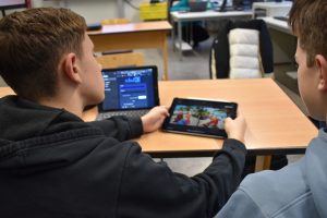 zwei Jugendliche sind von hinten zu sehen, sie sitzen an einem Schultisch. Ein Jugendlicher hält ein Tablet, auf dem KI generierte Bilder zu sehen sind. Die beiden Jugendlichen schauen auf den Bildschirm. Auf dem Tisch steht ein weiteres Tablet.